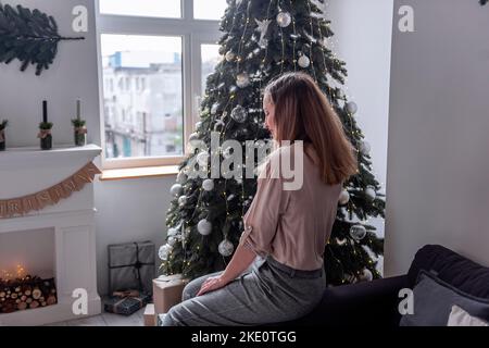 A stylish millennial woman with red lips, in a delicate shirt and gray trousers sits on a gray trendy sofa near the Christmas tree. Scandinavian inter Stock Photo