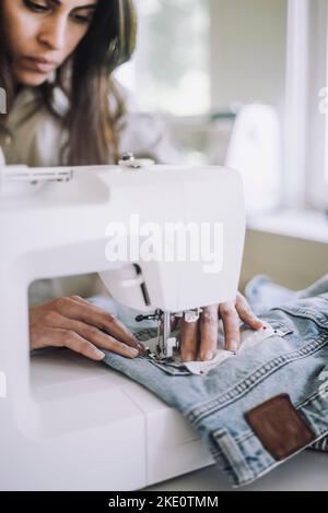 Female fashion designer sewing fabric on jeans pocket using sewing machine at workshop Stock Photo