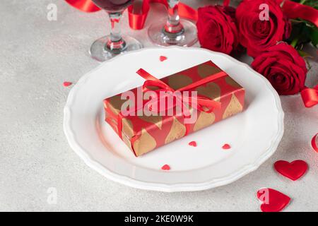 Gift in wrapping paper on white plate, red roses and two glasses for wine on light table, concept for Valentines Day Stock Photo