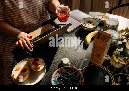 High angle view of woman holding drinking glass while using laptop with breakfast on table at home Stock Photo