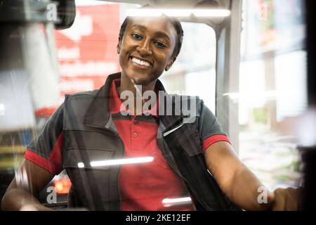Portrait of happy female warehouse worker sitting in forklift seen through windshield Stock Photo