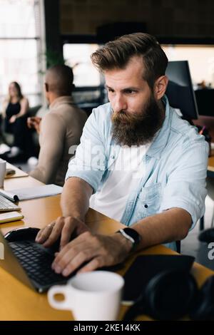 Bearded male computer programmer working on laptop at desk in office Stock Photo