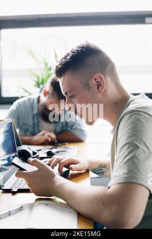 Male computer programmer using smart phone by laptop on desk in startup company Stock Photo