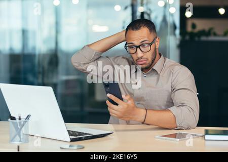 African american man looking and thinking on the phone, businessman working with laptop and computer in the middle of modern office building, young investor in glasses and shirt thinking. Stock Photo