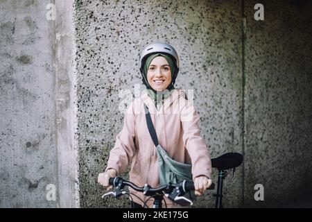 Portrait of smiling young woman with cycle standing near wall Stock Photo
