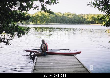 Man with paddleboard looking at lake while sitting on jetty Stock Photo