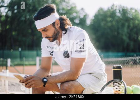 Man wearing headband using smart phone sitting on bench at tennis court Stock Photo