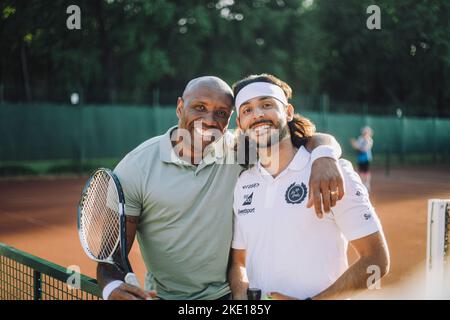 Happy man with arm around male friend wearing headband at tennis court Stock Photo