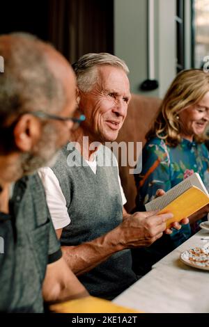 Senior man holding book while talking with male and female friends in cafe Stock Photo