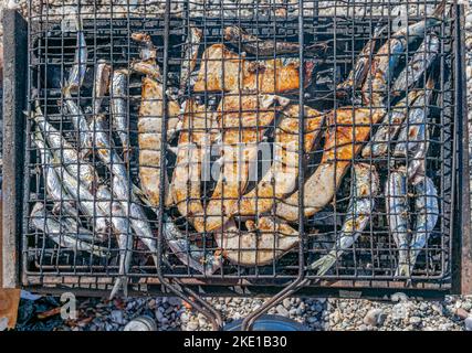 Overhead view of grilled slices of swordfish and sardines on a barbecue grill outdoors under sunlight. Blurred beach pebbles in background. Stock Photo