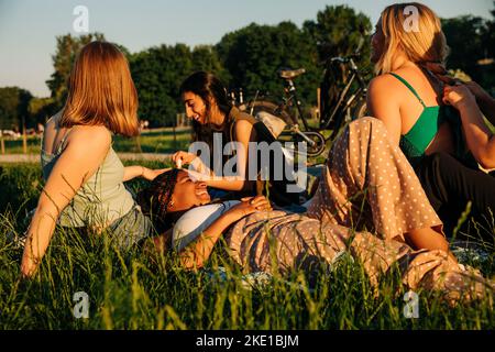 Smiling teenage girl lying on grass while talking with female friends at park during sunset Stock Photo