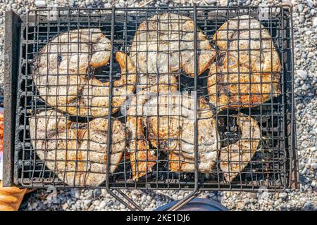 Overhead view of grilled slices of swordfish in a barbecue grill with a bread and blurred beach pebbles on background. Stock Photo