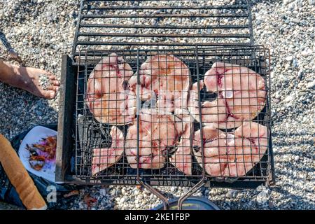 Overhead view of grilled slices of swordfish in a barbecue grill, a bread, a plate of shrimps and a foot with beach pebbles on blurred  background. Stock Photo
