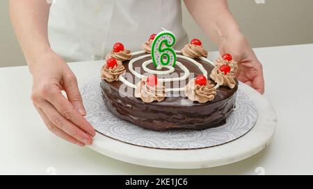 Woman decorating cake with candle in the form of number Six. Homemade cake decorated with chocolate cream and cherries close up Stock Photo