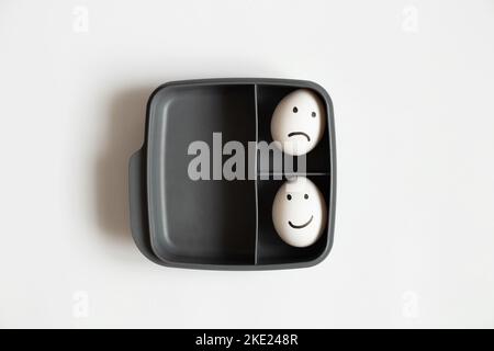 Two eggs with a joyful smile and a sad one lie in a plastic container on a white background, a choice of joy and sadness, lunch Stock Photo