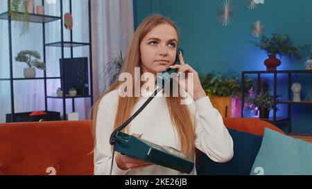 Crazy adult girl in casual shirt talking on wired vintage old-fashioned retro telephone of 80s, fooling, making silly faces. Young woman sitting on orange couch at home living room making conversation Stock Photo