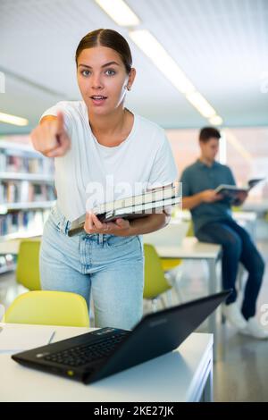 Serious young female student pointing finger into camera while staninding with the book in the library Stock Photo