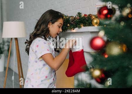 Side view of shocked child in pajama looking at Christmas stocking near fireplace at home Stock Photo