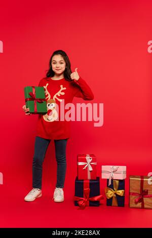 full length of happy child in knitted sweater showing thumb up near Christmas presents on red Stock Photo