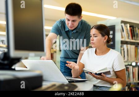 Man in casual clothes asking young lady to be more quiet while she is reading books in the library Stock Photo