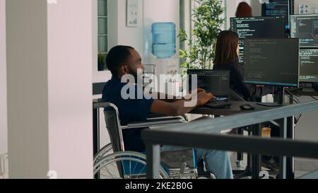 African american IT engineer with disability typing code in it development agency office, using terminal window to write html script database. Wheelchair user working with security programming. Stock Photo