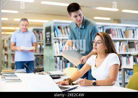 Young man bringing book to female in glasses writing in planner using laptop in the library Stock Photo