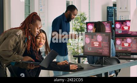 Diverse group of engineers having computer hacked and system crash on multiple monitors, reading critical error message flashing on screen. Security breach alert and hacking warning. Handheld shot. Stock Photo