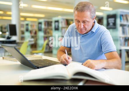 Focused casual mature male writing in notepad and reading books in the library Stock Photo