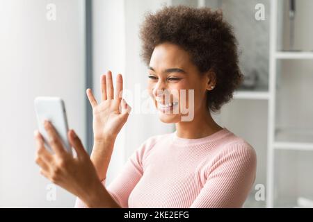 Smiling young african american female chatting on phone, has video call, waving hand at webcam Stock Photo