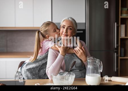 Glad happy caucasian little girl hugs and kisses in cheek of elderly grandmother, prepares dough for baking Stock Photo