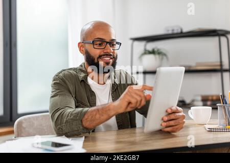 Modern technologies. Happy mature latin man using digital tablet, sitting at desk, browsing internet on modern gadget Stock Photo
