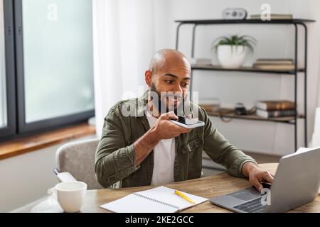 Positive latin business man recording audio message on smartphone and using laptop computer, working at home Stock Photo