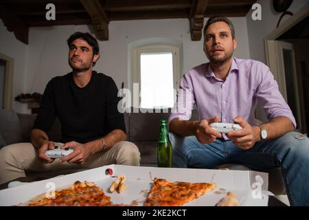 Two young happy friends playing video games while sitting on the couch. Stock Photo