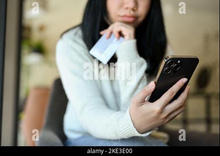 Thoughtful young Asian woman in casual clothes using mobile banking on her smartphone to transfer money or pay bills. credit card payment concept. cro Stock Photo