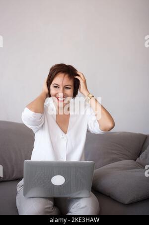 Adorable successful happy confident brunette woman 35-40 years old sits on the couch and works using a laptop. Modern technologies of communication, l Stock Photo