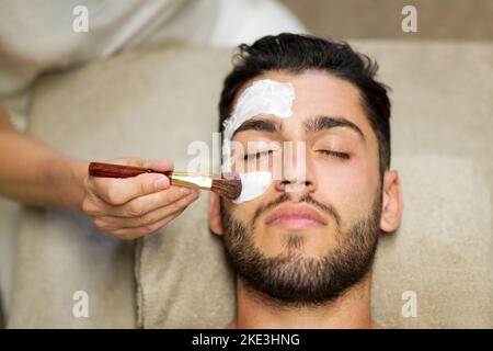 From above crop beautician applying clay mask on face of calm handsome bearded man lying on table in spa salon Stock Photo