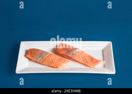 Two raw salmon loins seasoned with salt and pepper on a rectangular white ceramic platter on a blue studio background with copy space. Food and fish. Stock Photo