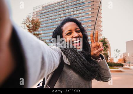 Carefree woman looking at camera and smiling while taking a selfie and showing peace sign outdoors. Lifestyle Concept. Stock Photo