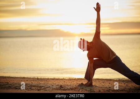 Challenge the body, quieten the mind. a man practicing the triangle pose during his yoga routine at the beach. Stock Photo