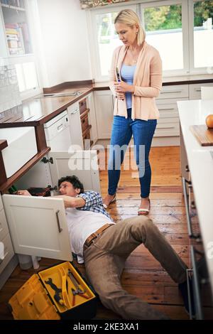 He knows a thing or two about plumbing. a man fixing pipes under his kitchen sink while his wife watches. Stock Photo