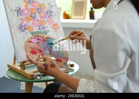 Cropped image of female artist holding palette and painting picture on canvas with oil paints in cozy art studio Stock Photo