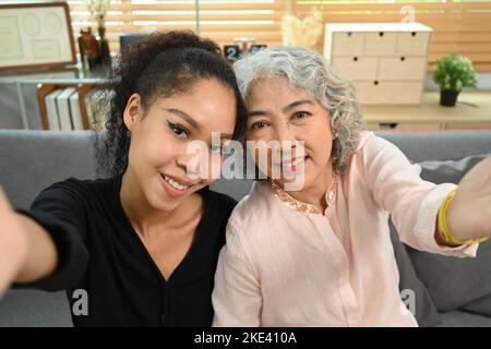 Happy elderly woman and grown up daughter having conversation by video call, taking selfie while sitting in cozy living room Stock Photo