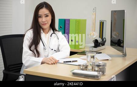 Portrait Of Smiling Asian Female Doctor Wearing White Coat With Stethoscope Sitting Behind Desk In Office. Stock Photo