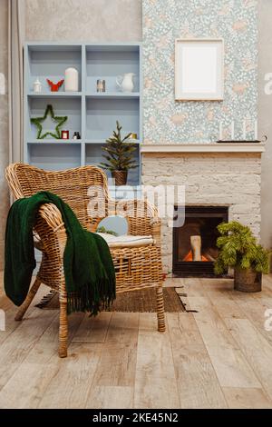 Rattan wicker chair and fireplace in the living room with Christmas decorations. Blank wooden frame on the wall. Shelving. Stock Photo