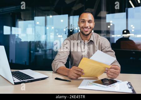 Portrait of happy african american man in office doing paperwork, man looking at camera and smiling holding good news notification bank letter. Stock Photo
