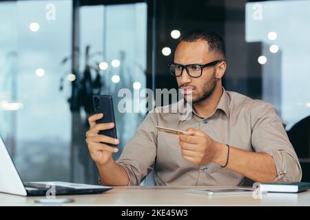 Dissatisfied and angry african american businessman inside office trying to make a bank transfer, man got rejected error, worker sitting at laptop holding bank credit card and phone. Stock Photo