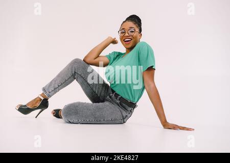 Cheerful African American woman sitting on the floor and smiling on gray background. Studio shot Stock Photo