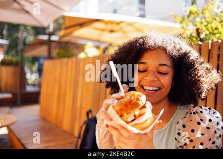 Young african american girl eating classic burger and drinking lemonade in the city. Outdoor woman wearing summer blouse with polka dots eatting delic Stock Photo