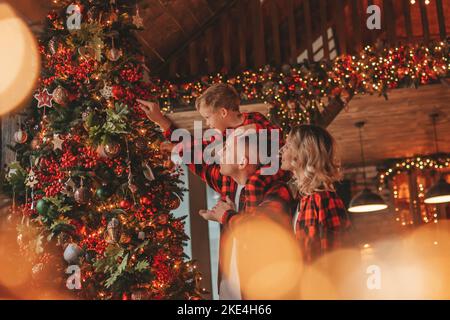 Smile active parents with small son in red checkered sleepwears waiting Santa indoor. Stylish family love laughing hugs kisses at home. Celebrate New Stock Photo