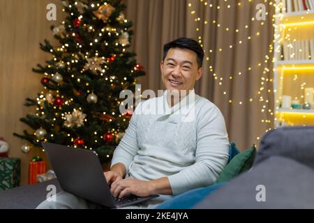 Festive portrait of young handsome man Asian freelancer businessman. Sitting at home on the sofa near the Christmas tree, holding a laptop, looking at the camera, smiling, in a good Christmas mood. Stock Photo